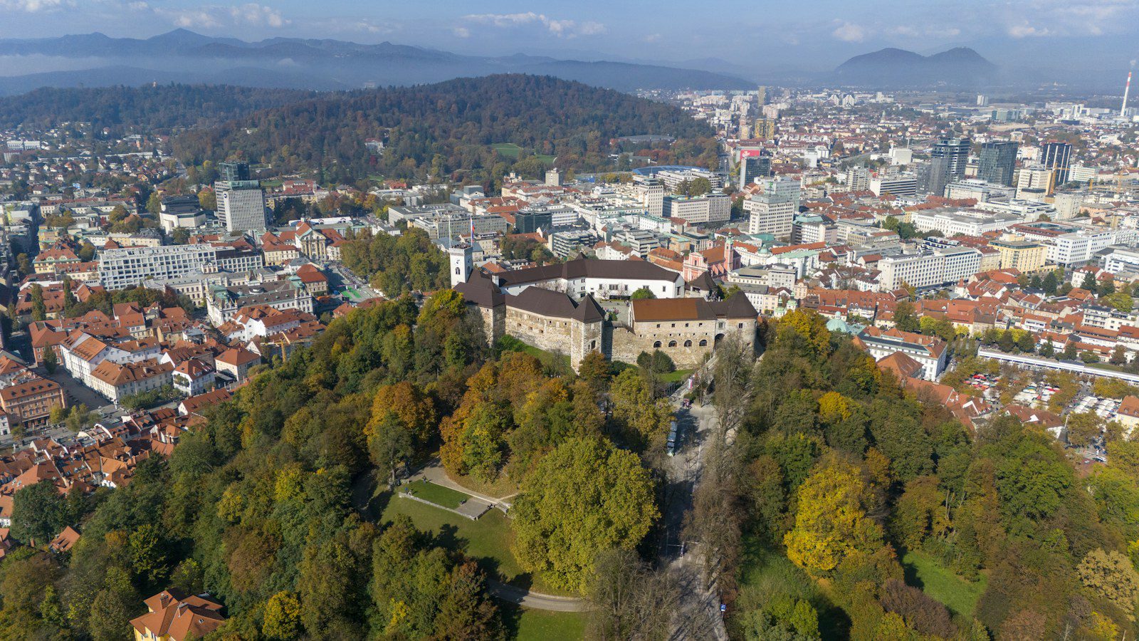 Lubiana, Slovenia: guida city break tra Castello, Ljubljanica e Ponti di Plečnik 2 Veduta aerea del Castello di Lubiana sulla collina boscosa con i colori dell’autunno, i tetti rossi del centro storico e le montagne sullo sfondo