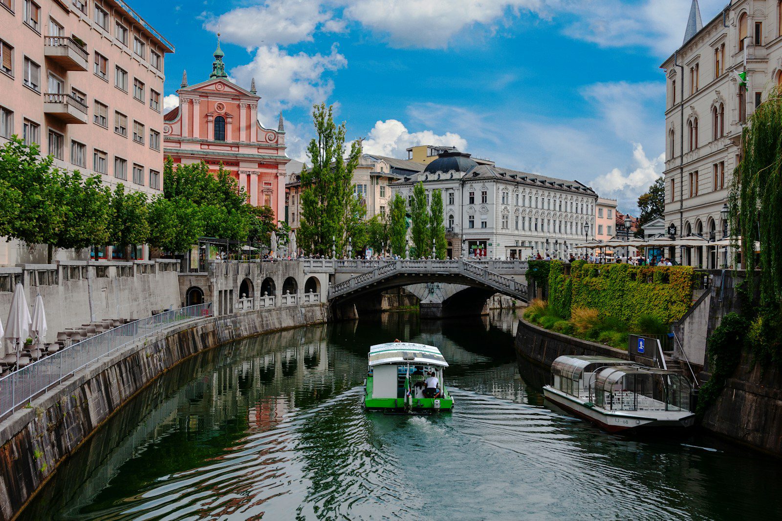 Battello turistico sulla Ljubljanica nel centro di Lubiana con il Ponte Triplo, la facciata rosa della Chiesa Francescana e i palazzi storici sul lungofiume