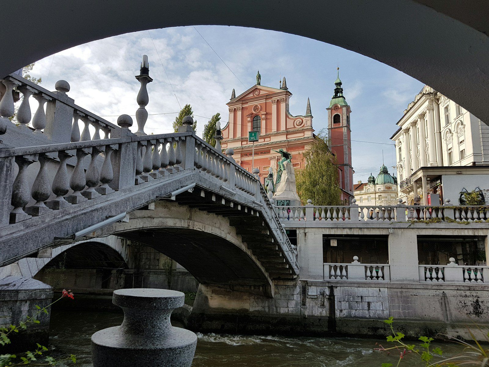 Lubiana, Slovenia: guida city break tra Castello, Ljubljanica e Ponti di Plečnik 1 Il Ponte Triplo di Lubiana visto da sotto un’arcata, con le balaustre di Plečnik, il monumento a Prešeren e la facciata rosa della Chiesa Francescana dell’Annunciazione